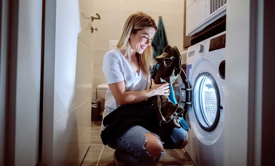 Vrouw knielt voor wasmachine in badkamer, met kleding in haar handen. Op de achtergrond zijn wastafel en handdoek te zien.