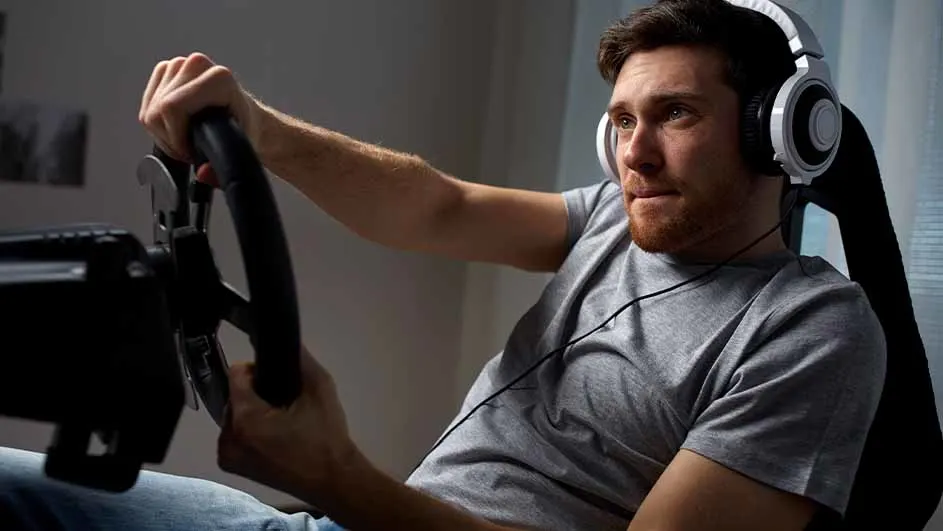 Een man met een grijze shirt en witte hoofdtelefoon houdt een zwart stuur vast in een donkere kamer.