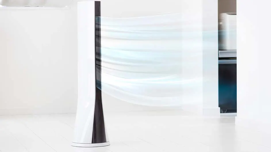 Een witte, verticale ventilator met zwarte accenten blaast lucht in een witte kamer. Op de achtergrond staat een kast.