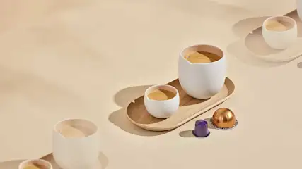 Twee koffiekopjes met romige espresso staan op een houten dienblad, naast een paar koffiecapsules op een lichte achtergrond.