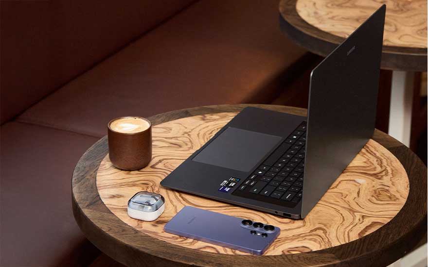 Café setting met ronde houten tafels, een open Samsung laptop, paarse smartphone, oplaadcase voor oordopjes en een cappuccino met romige schuimlaag.