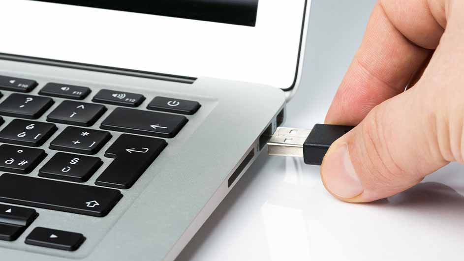 Een hand die een zwarte USB-stick in de poort van een grijze laptop steekt, met een wit oppervlak eronder.
