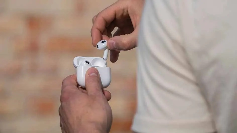 Someone is taking a wireless earbud out of its white charging case, with hands clearly visible in the foreground of the photo.