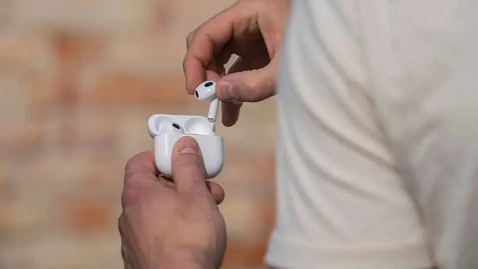 Someone is taking a wireless earbud out of its white charging case, with hands clearly visible in the foreground of the photo.