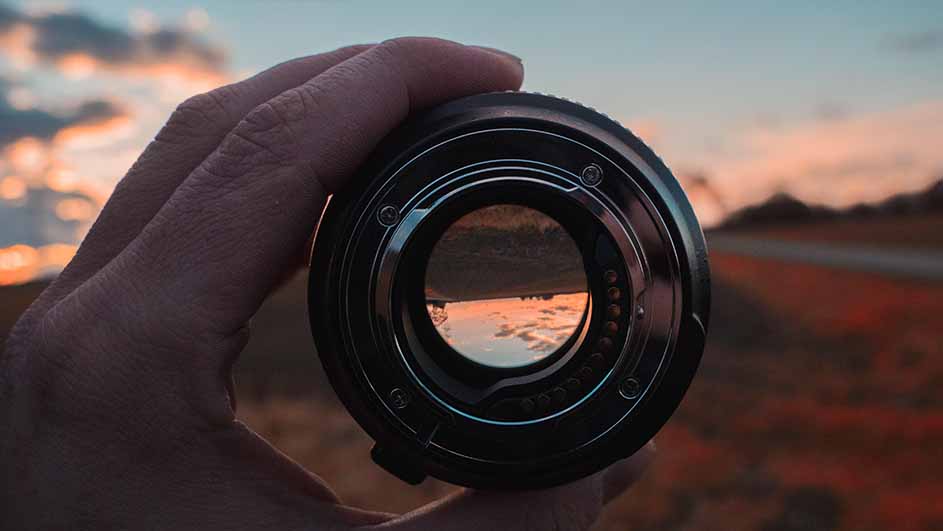 Een hand houdt een cameralens vast, met een zonsondergang en landschap zichtbaar door de lens. De lucht is blauw en oranje.