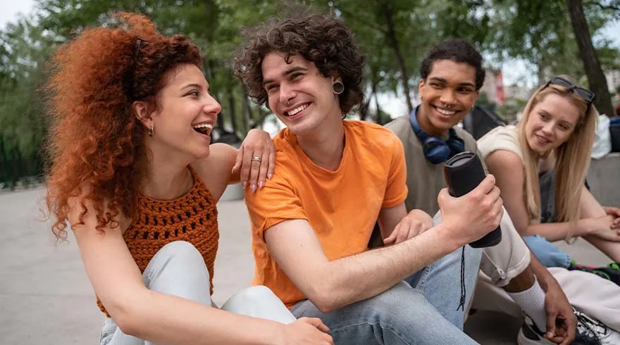 Vier lachende jongeren zitten buiten met een zwarte speaker. De achtergrond toont bomen en vage gebouwen.