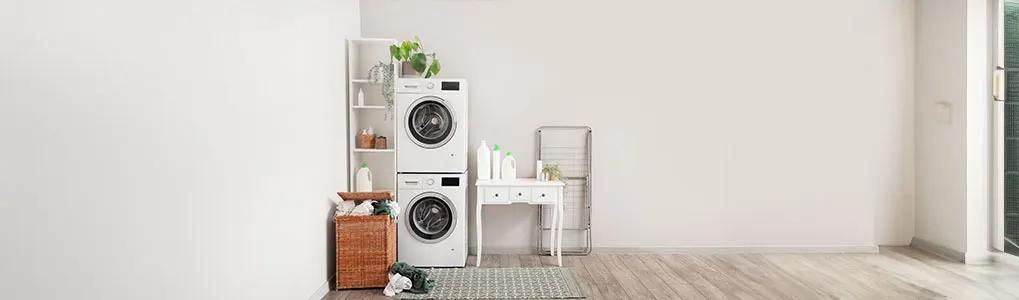 A laundry room features a stacked washer and dryer next to a basket with clothes, with detergent on a table.