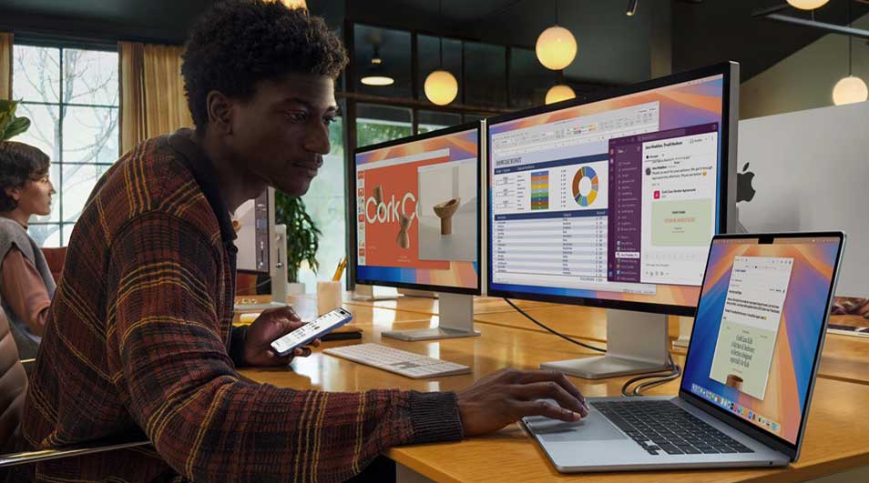 Young man in a modern office uses a laptop and dual monitors showing charts, chat and slides, while holding his smartphone nearby.