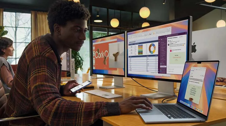 Young man in a modern office uses a laptop and dual monitors showing charts, chat and slides, while holding his smartphone nearby.