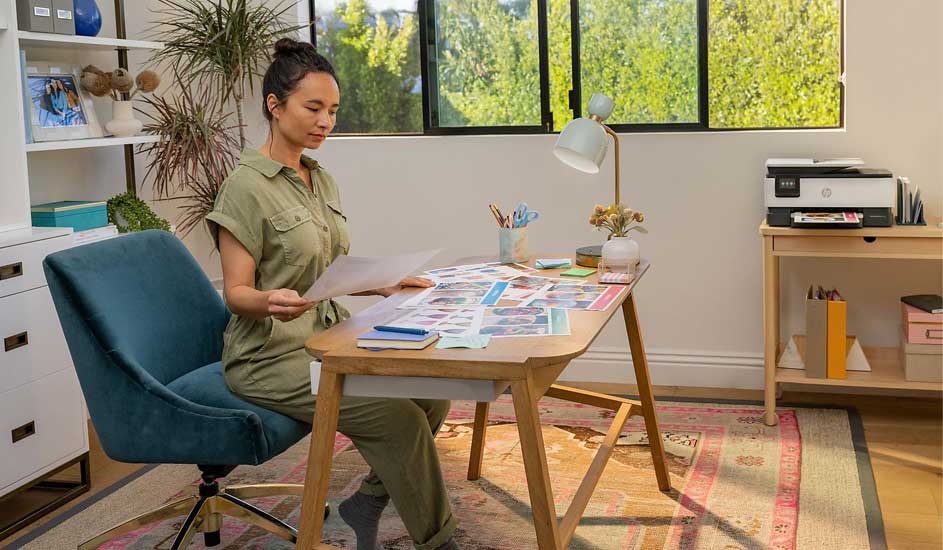 Een vrouw zit aan een bureau in een lichte kamer en bekijkt papieren. Op de achtergrond staat een HP printer op een houten tafel. Merknaam: HP.