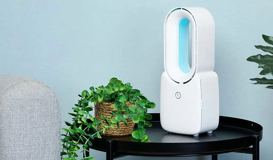 A Duux fan with blue light is on a black table beside a plant and grey sofa in a modern room.