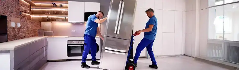 Two workers in blue uniforms move a stainless steel refrigerator on a dolly in a kitchen.