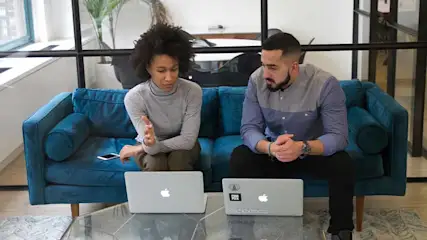 Een vrouw en een man zitten naast elkaar op een blauwe bank, allebei werkend op een Apple-laptop. De vrouw maakt een handgebaar terwijl ze praat, de man luistert aandachtig. Op het glazen tafeltje voor hen ligt een smartphone. Ze bevinden zich in een moderne kantoorruimte met glazen wanden.
