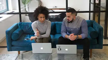 Two people sit on a blue sofa with laptops on a glass table. The background includes a window and indoor plants.