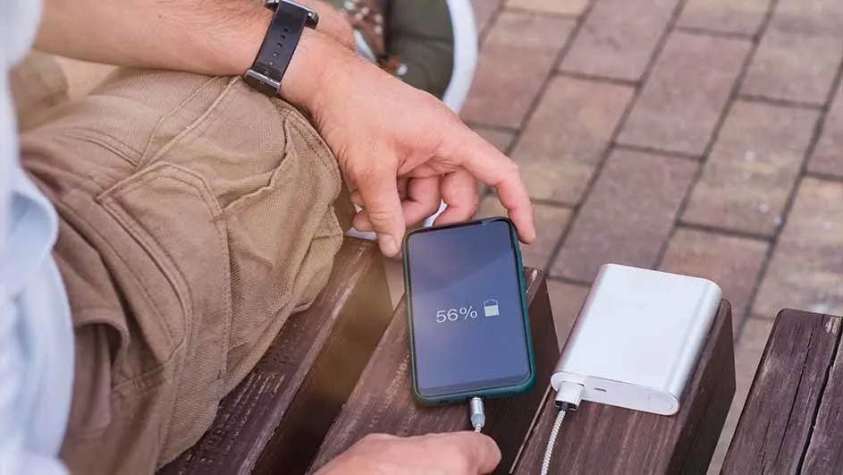 Een persoon zit op een bank en laadt een telefoon op met een witte powerbank. Het telefoonscherm toont 56% batterij.