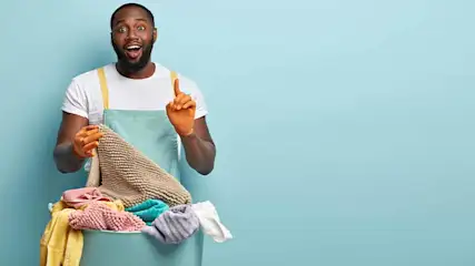 Een man met oranje handschoenen en een blauw schort houdt een wasmand vol handdoeken vast tegen een lichtblauwe achtergrond.