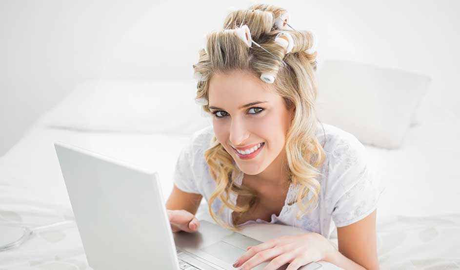 A smiling blonde woman with hair rollers lies on a bed and uses a laptop in a bright room.