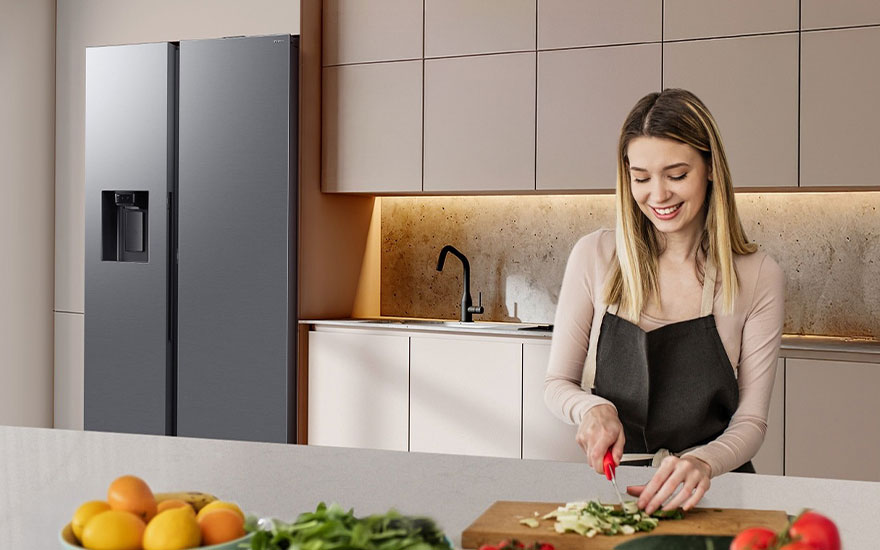 Vrouw kookt in moderne keuken met grijze koelkast, verse groenten op werkblad, warme verlichting en neutrale tinten.