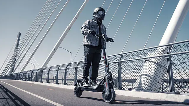 Een persoon met een helm rijdt op een elektrische scooter op een brug met witte kabels tegen een blauwe hemel.