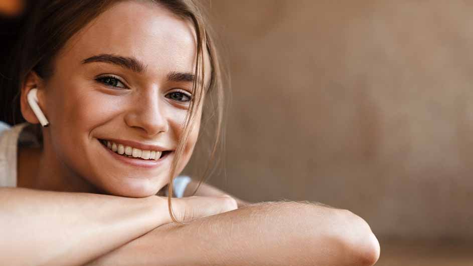 Jonge vrouw glimlacht met een witte draadloze oortelefoon. Haar armen zijn gekruist en haar hoofd rust erop.