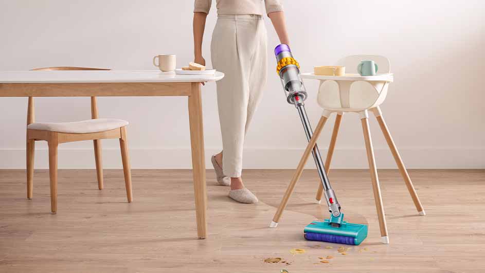 Vrouw stofzuigt naast een houten tafel en een kinderstoel op een houten vloer met kruimels.