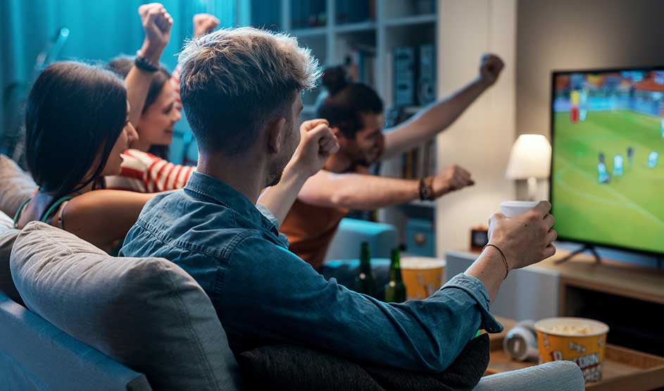 Vrienden kijken samen naar voetbal op televisie met enthousiasme en gebaren. Man op bank in denim shirt met drankje, anderen juichen. Gezellige, feestelijke thuisbioscoopsfeer met snacks.