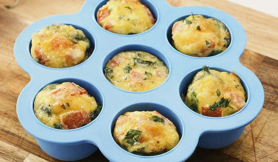 In een blauwe siliconen bakvorm liggen zes hartige muffins met kaas, tomaat en kruiden op een houten snijplank. Merk is niet zichtbaar.