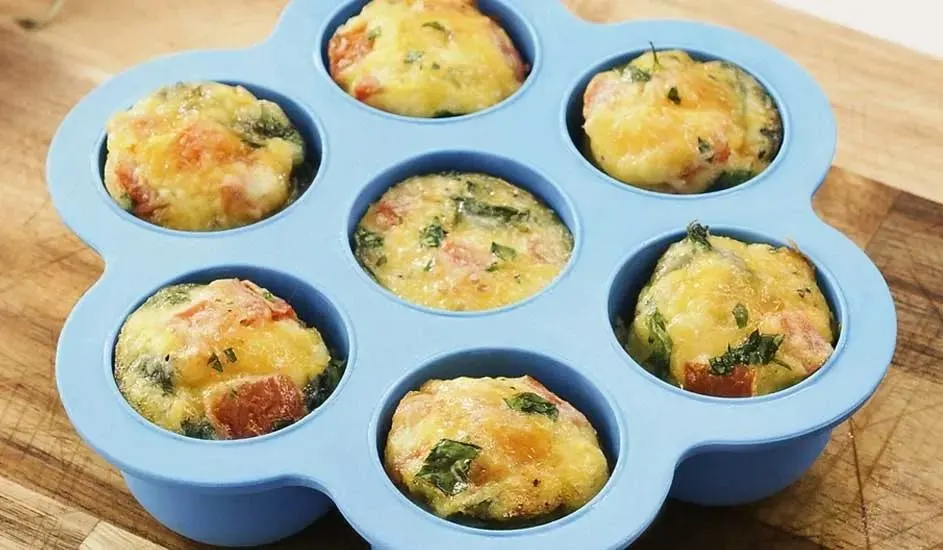 In een blauwe siliconen bakvorm liggen zes hartige muffins met kaas, tomaat en kruiden op een houten snijplank. Merk is niet zichtbaar.