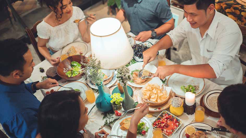 Een groep mensen zit aan tafel, vol met borden, salades, fruit en drankjes, onder een lamp.