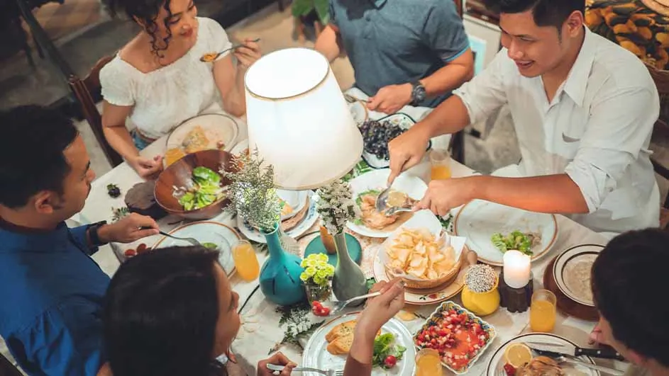 Een groep mensen zit aan tafel, vol met borden, salades, fruit en drankjes, onder een lamp.