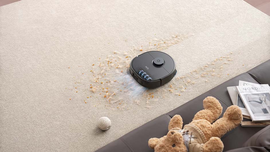 Een ronde, zwarte robotstofzuiger reinigt een beige vloerkleed met vuil. Een bal, knuffel en tijdschriften liggen op een grijze bank.