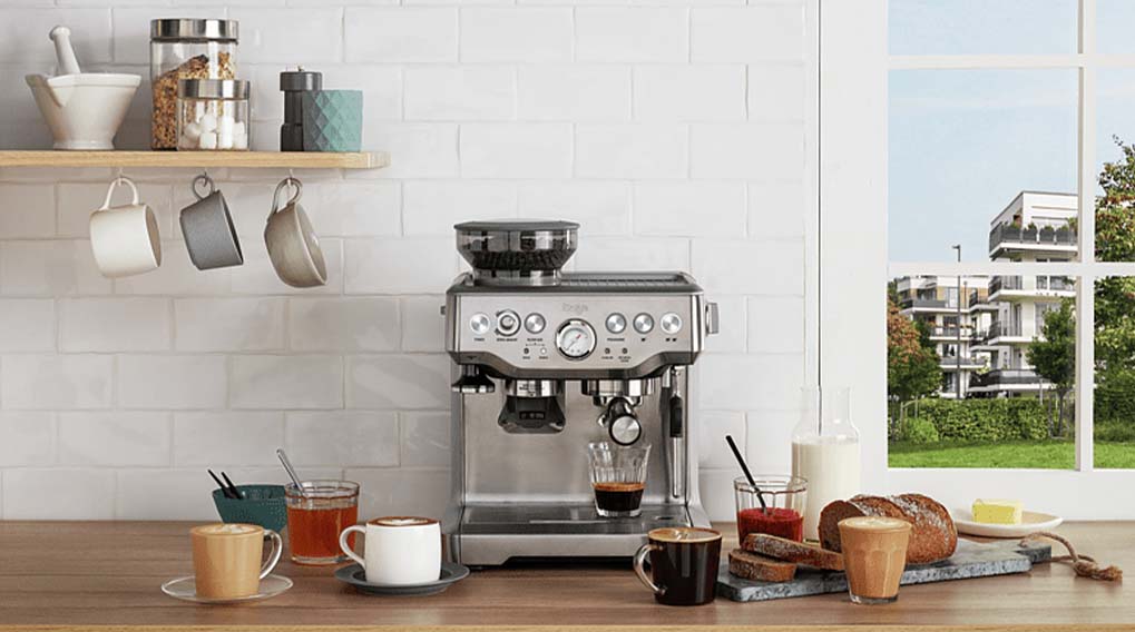 Een zilveren espressomachine op een houten aanrecht met kopjes, brood en een blik op een gebouw door het raam.