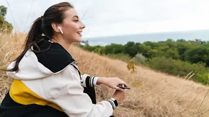 Een vrouw met oortjes en een hoodie zit in hoog gras. Op de achtergrond zijn bomen en de zee te zien.