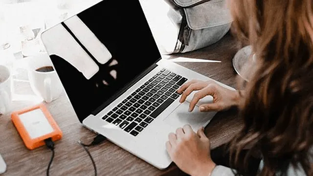 Vrouw typt op een laptop op een houten bureau. Er staat een oranje harde schijf, koffie en een grijze tas op het bureau.