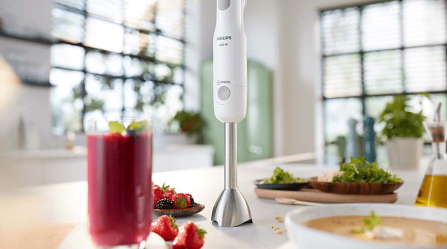 Een keukenopstelling met een witte staafmixer, rode smoothie, aardbeien, salade en soep tegen een lichte achtergrond.