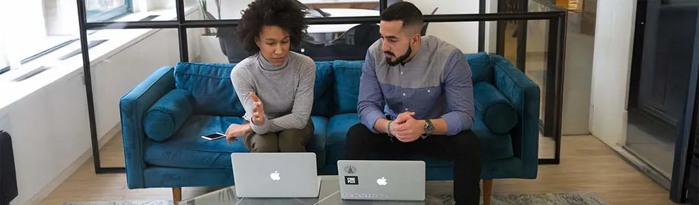 Een vrouw en een man zitten naast elkaar op een blauwe bank, allebei werkend op een Apple-laptop. De vrouw maakt een handgebaar terwijl ze praat, de man luistert aandachtig. Op het glazen tafeltje voor hen ligt een smartphone. Ze bevinden zich in een moderne kantoorruimte met glazen wanden.