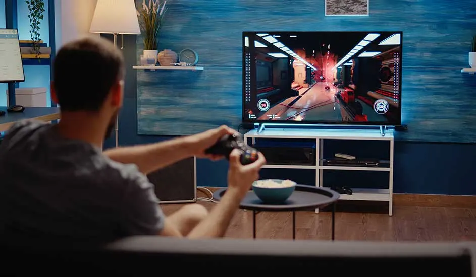 Een man speelt een videogame met een zwarte Xbox-controller, zittend op de bank en gericht op een groot tv-scherm in een gezellige kamer.