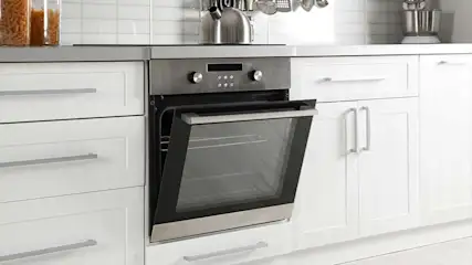A kitchen features white cabinets, a stainless steel oven with its door ajar, and assorted utensils hanging above.