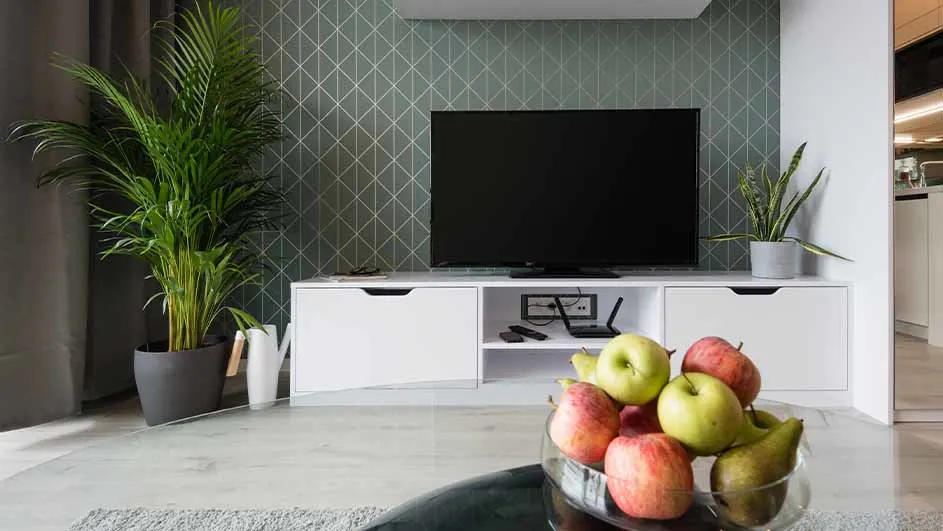 Woonkamer met een tv op een wit dressoir, fruit in een glazen schaal, planten en een grijs geometrisch behang.