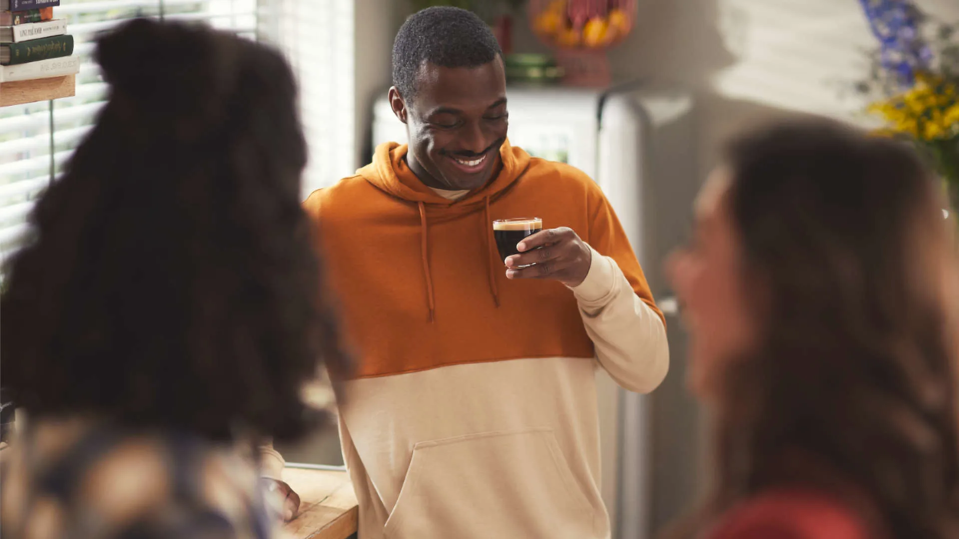 Een lachende man met een oranje-wit sweatshirt houdt een glas donkere drank vast in een keuken met twee andere mensen.
