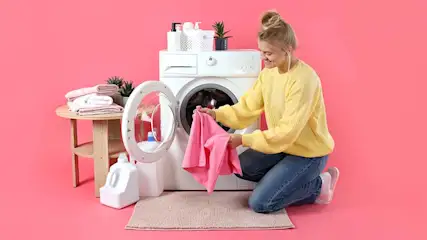 Vrouw knielt voor een wasmachine met een open deur en houdt een roze handdoek vast. Ernaast staat een tafeltje met handdoeken en plant.