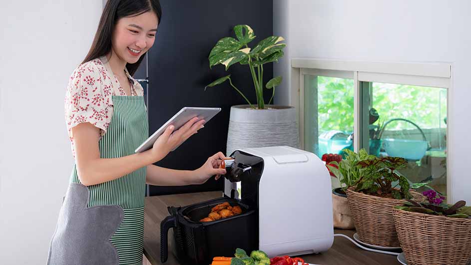 Een vrouw met een tablet bedient een witte airfryer in een keuken met planten en groenten.