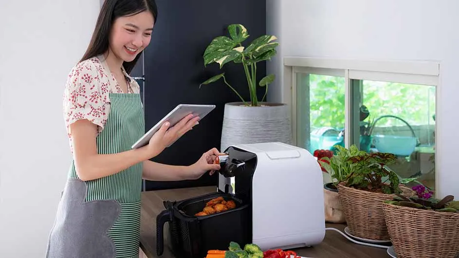Een vrouw met een tablet bedient een witte airfryer in een keuken met planten en groenten.