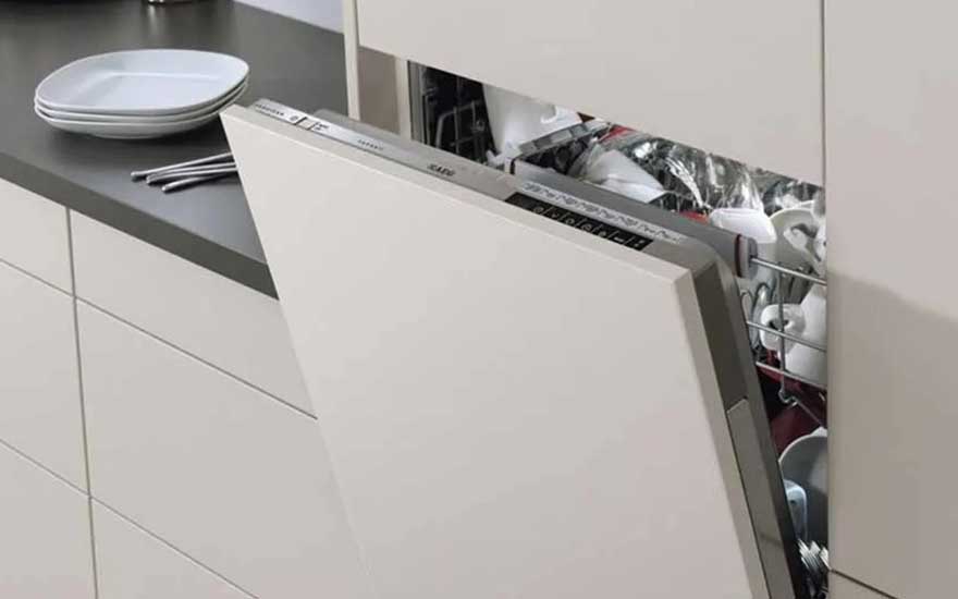 An AEG dishwasher, built into sleek white cabinets, is open and filled with clean dishes; plates and cutlery are placed on the dark worktop above.