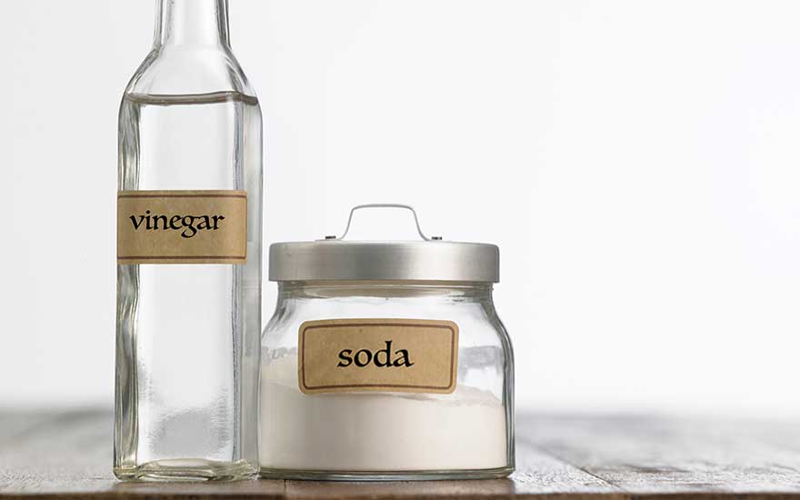 A glass bottle with a brown label reading "vinegar", partially filled with clear liquid. Beside it stands a glass jar with a metal lid, containing white powder and labelled "soda". Both are positioned on a light-coloured surface.
