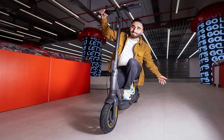 Young man cheerfully rides an electric scooter through a futuristic shop interior with red features and glowing signage.