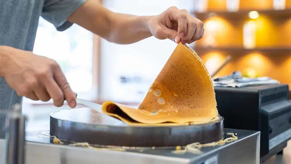 Een persoon maakt een crêpe op een ronde kookplaat met een spatel. Achtergrond: vage plank met flessen.