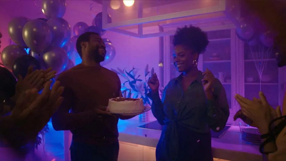 Mensen vieren feest met ballonnen en een taart. Een man draagt een taart; een vrouw danst.