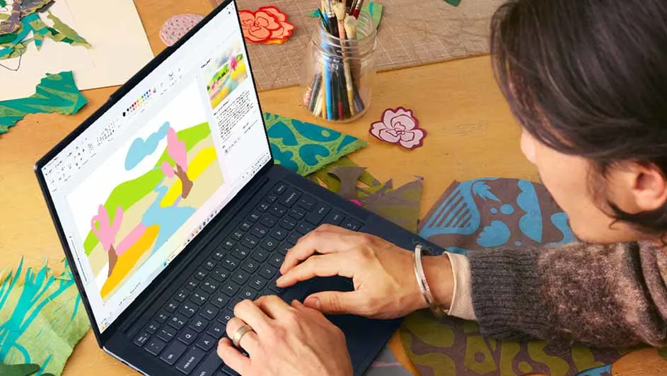 Een vrouw typt op een laptop met een tekening op het scherm. Penselen in een pot en bloemdecoraties staan op een tafel.