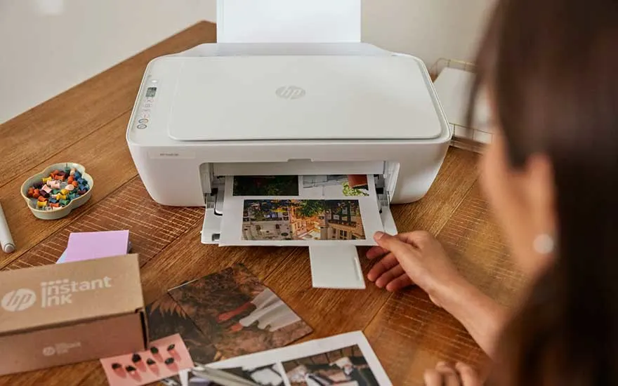 A white printer on a wooden desk is printing colourful photos. Nearby lie photos, ink cartridges and some stationery.